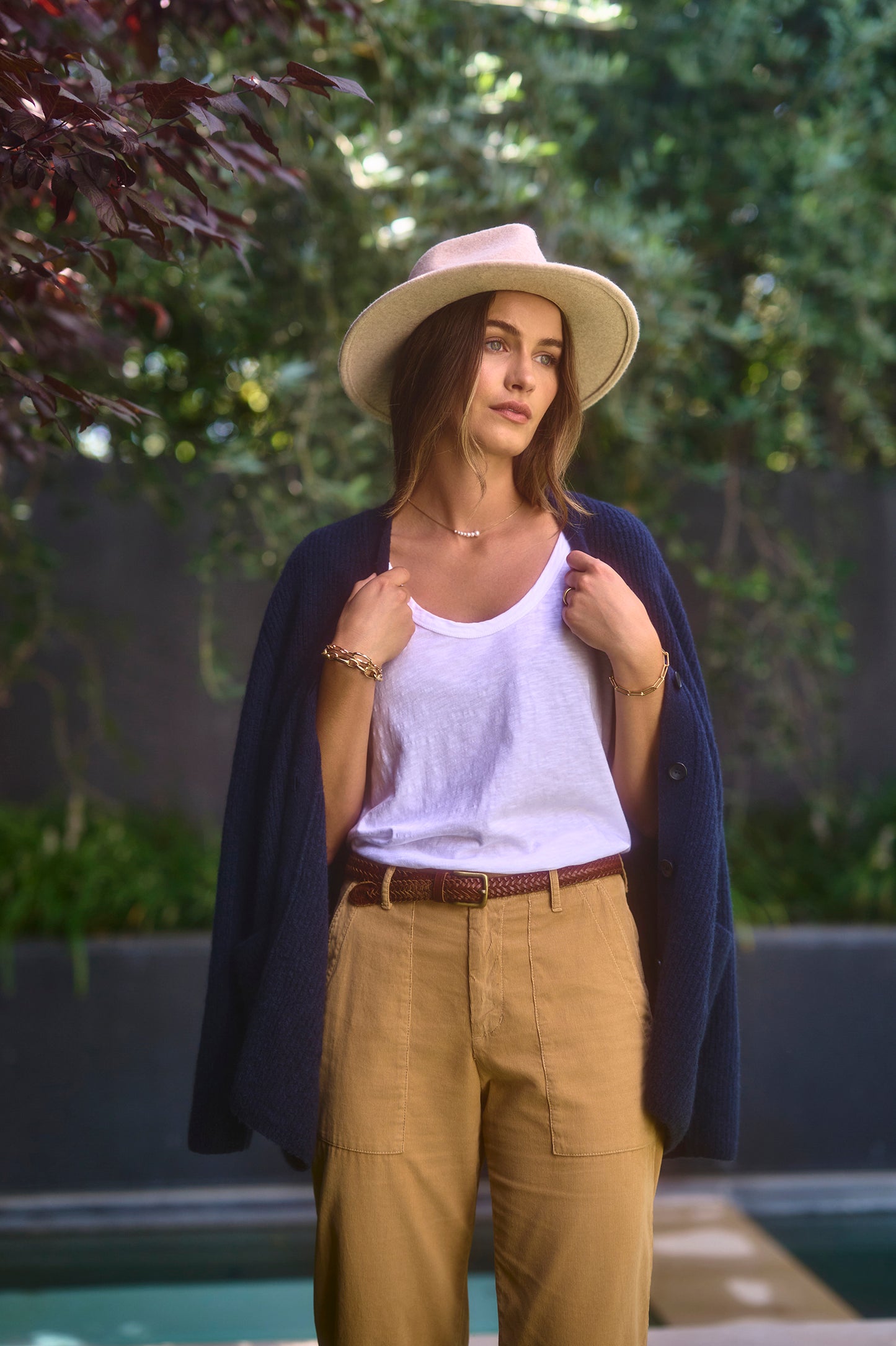A woman stands outdoors wearing the KIRA TEE by Velvet by Graham & Spencer, a white tank top made of cotton slub fabric. She matches it with brown pants, a beige hat, and has a blue sweater draped over her shoulders while holding onto it. Greenery serves as the backdrop.-38116478288065
