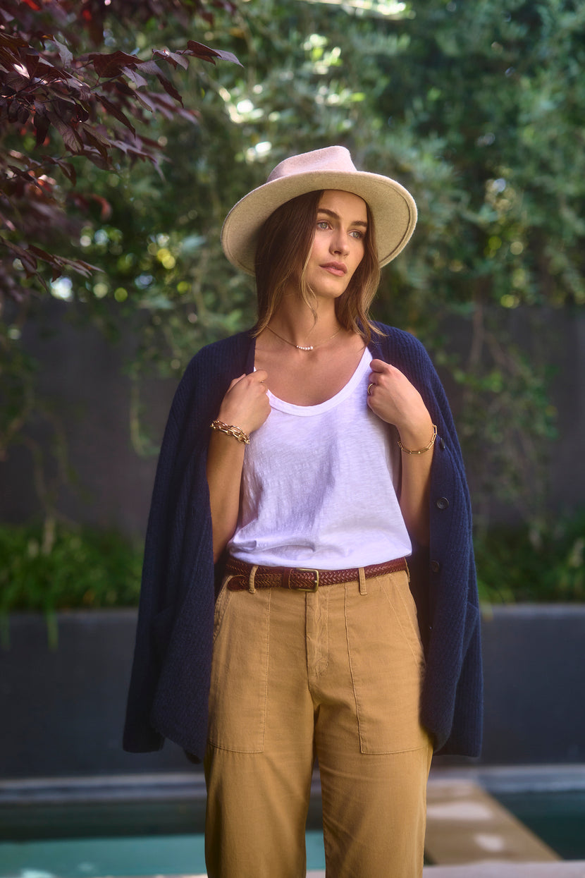 A woman stands outdoors wearing the KIRA TEE by Velvet by Graham & Spencer, a white tank top made of cotton slub fabric. She matches it with brown pants, a beige hat, and has a blue sweater draped over her shoulders while holding onto it. Greenery serves as the backdrop.