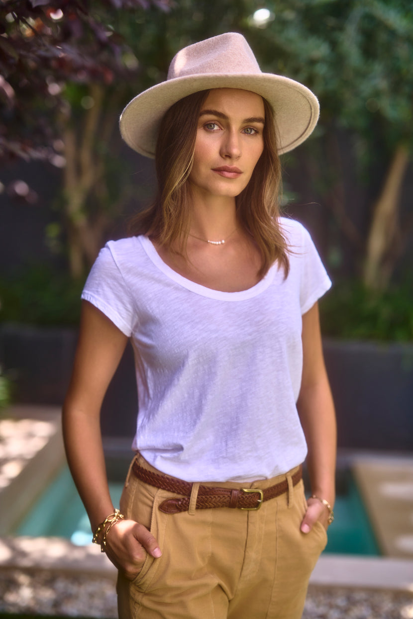 A woman stands outdoors, wearing the KIRA TEE by Velvet by Graham & Spencer and light brown pants. She has a beige hat on and her hands in her pockets, while greenery fills the background.