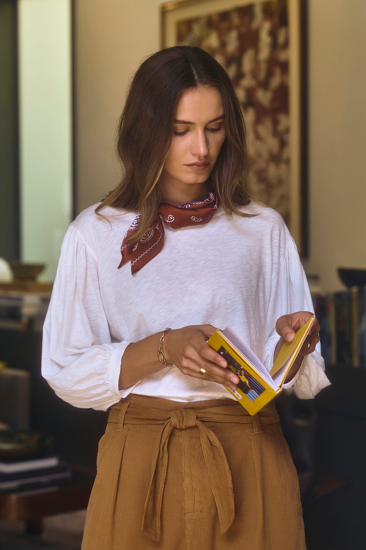 A person in a white AMARA TEE with banded cuffs by Velvet by Graham & Spencer and brown pants reads a small yellow book, standing indoors.-38139242545345