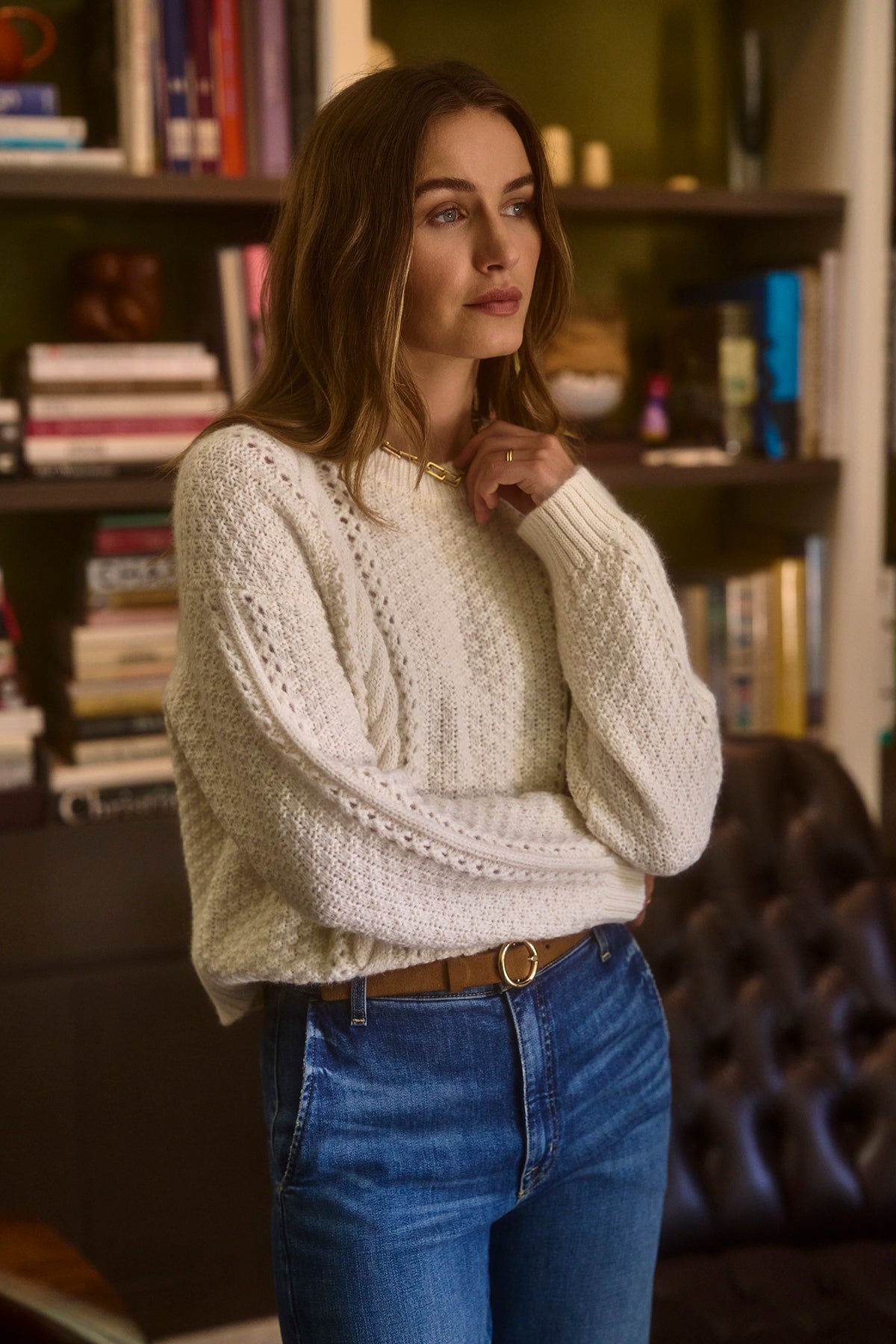   A person wearing the MARLEY SWEATER by Velvet by Graham & Spencer, which is a white, cable-knit alpaca blend, paired with jeans stands indoors with bookshelves in the background. 