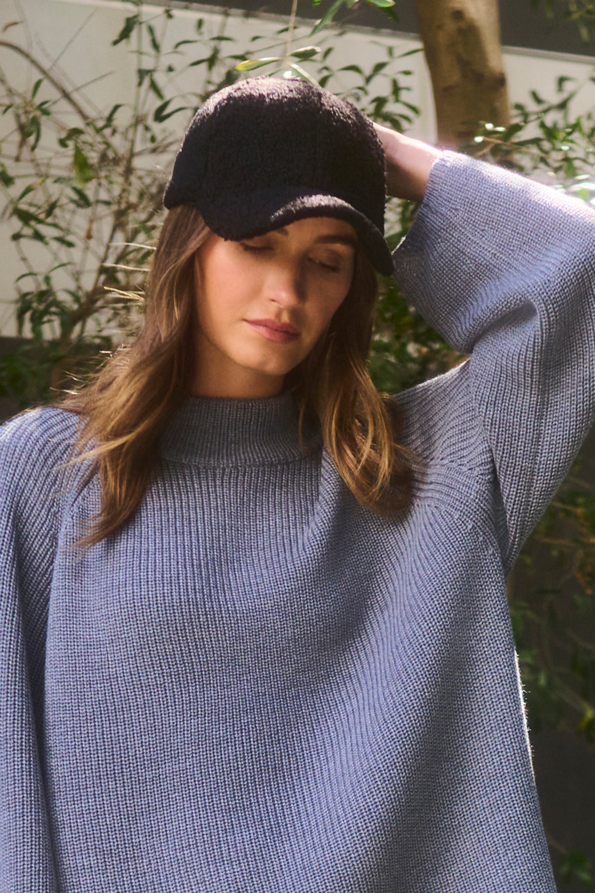   A woman wearing the SHERPA CAP by Velvet by Graham & Spencer, along with an eco faux-sherpa gray sweater, stands outdoors with her eyes closed and one hand behind her head. 