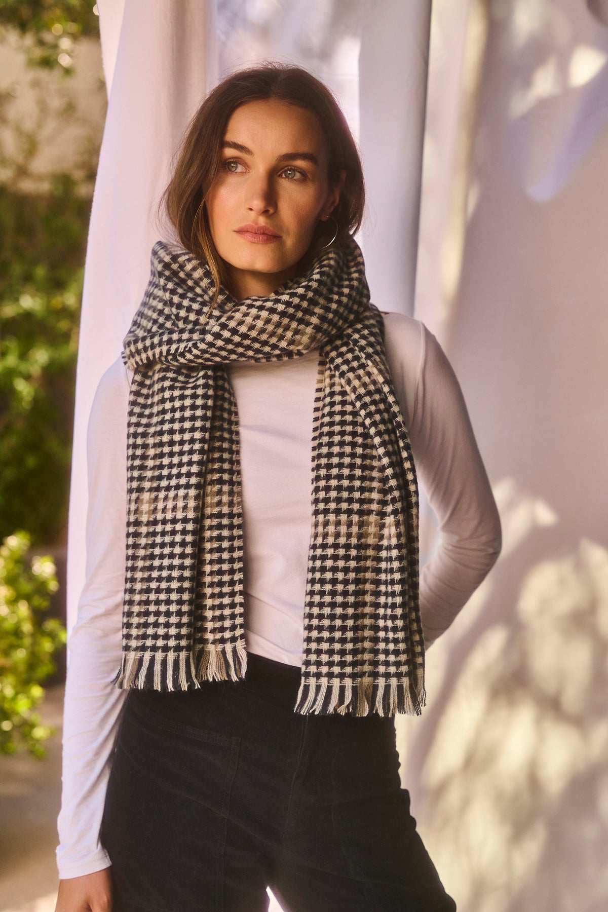 A woman stands indoors wearing a white long-sleeve shirt, black pants, and the PEAK SCARF by Velvet by Graham & Spencer in a black-and-white houndstooth pattern.-37930200170689