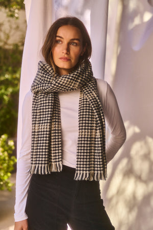 A woman stands indoors wearing a white long-sleeve shirt, black pants, and the PEAK SCARF by Velvet by Graham & Spencer in a black-and-white houndstooth pattern.