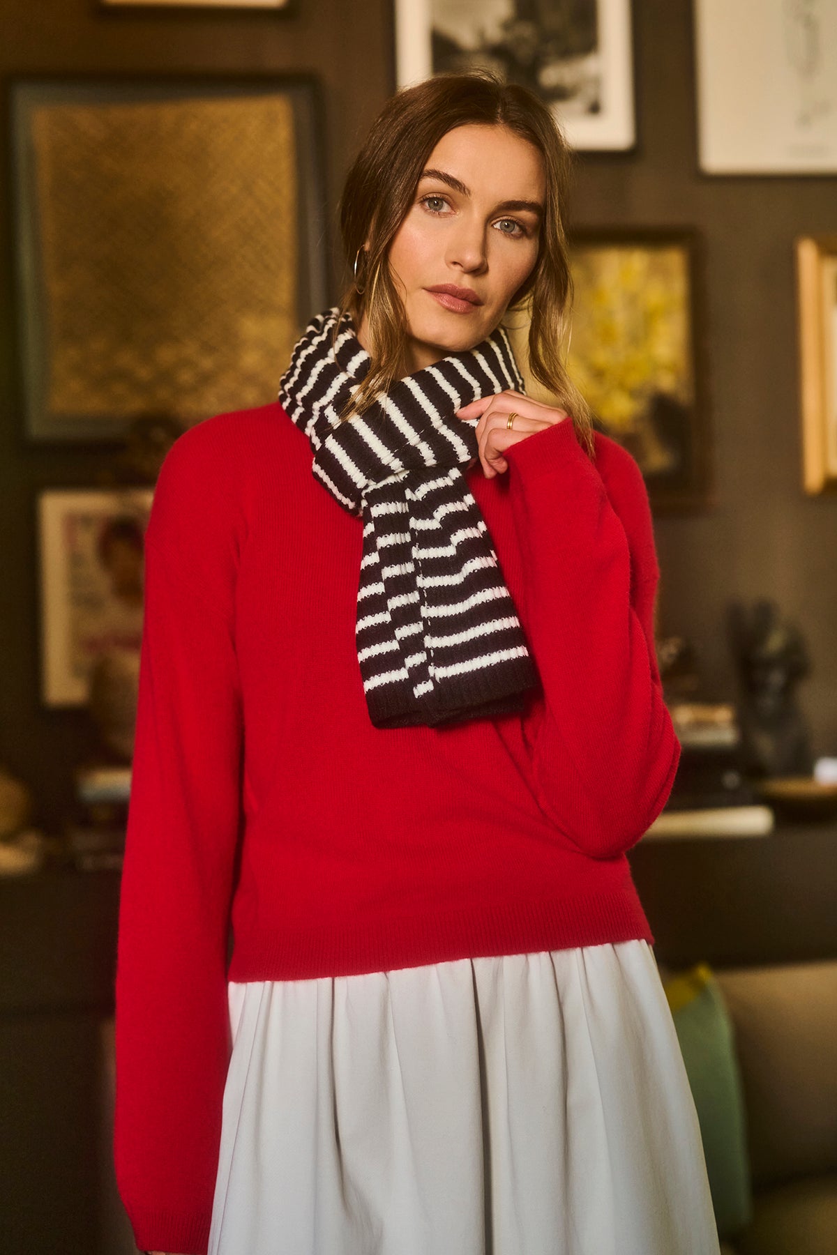   A person wearing a red sweater, the BOLTON STRIPE SCARF by Velvet by Graham & Spencer, and a white skirt, posing indoors with framed artwork in the background. 