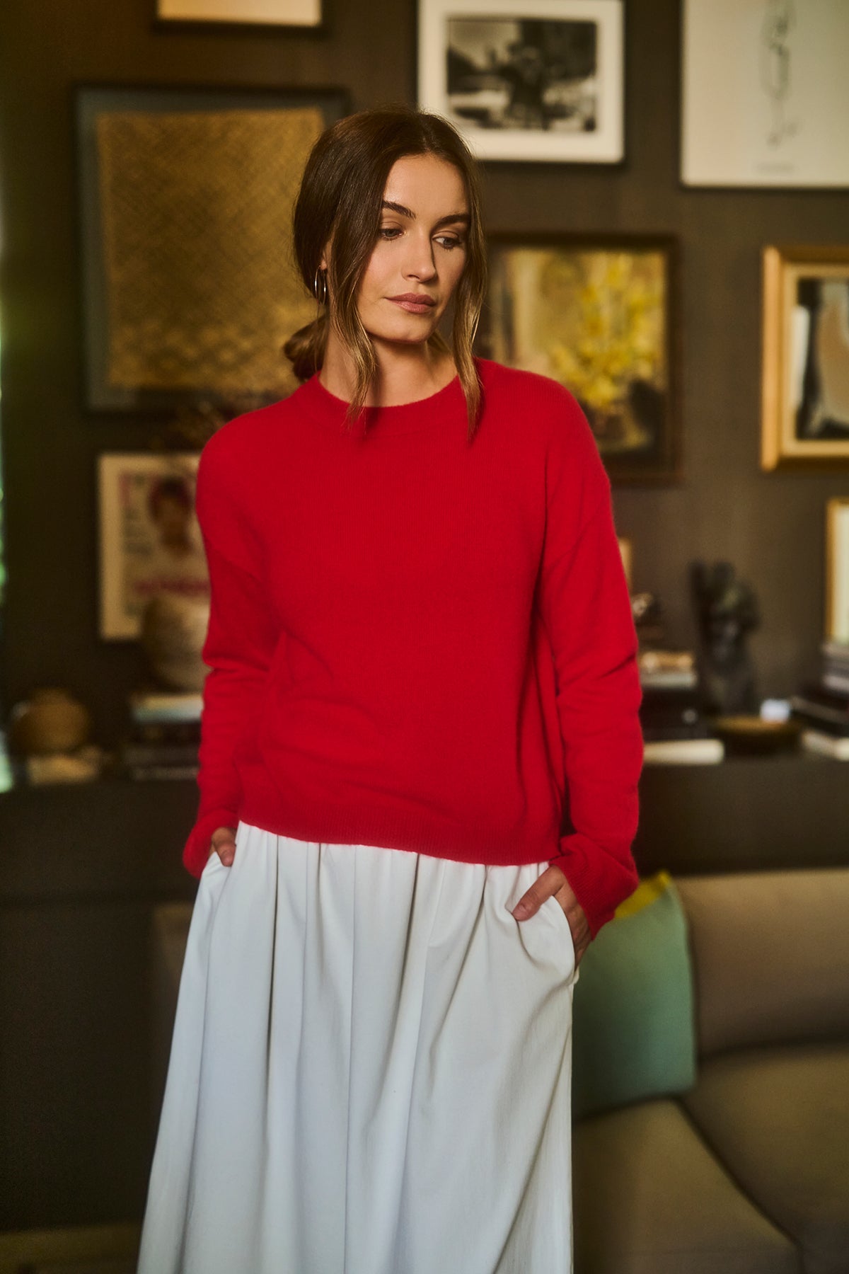   A person in a red BROOKLYN CASHMERE SWEATER by Velvet by Graham & Spencer with a crew neckline and white skirt stands in a room adorned with framed pictures on the wall. 