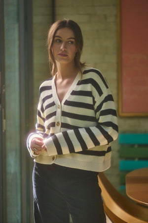 A woman in the ELODIE CARDIGAN by Velvet by Graham & Spencer, featuring a cotton cashmere blend with striped details, stands indoors near large windows with sunlight casting a gentle glow around her.