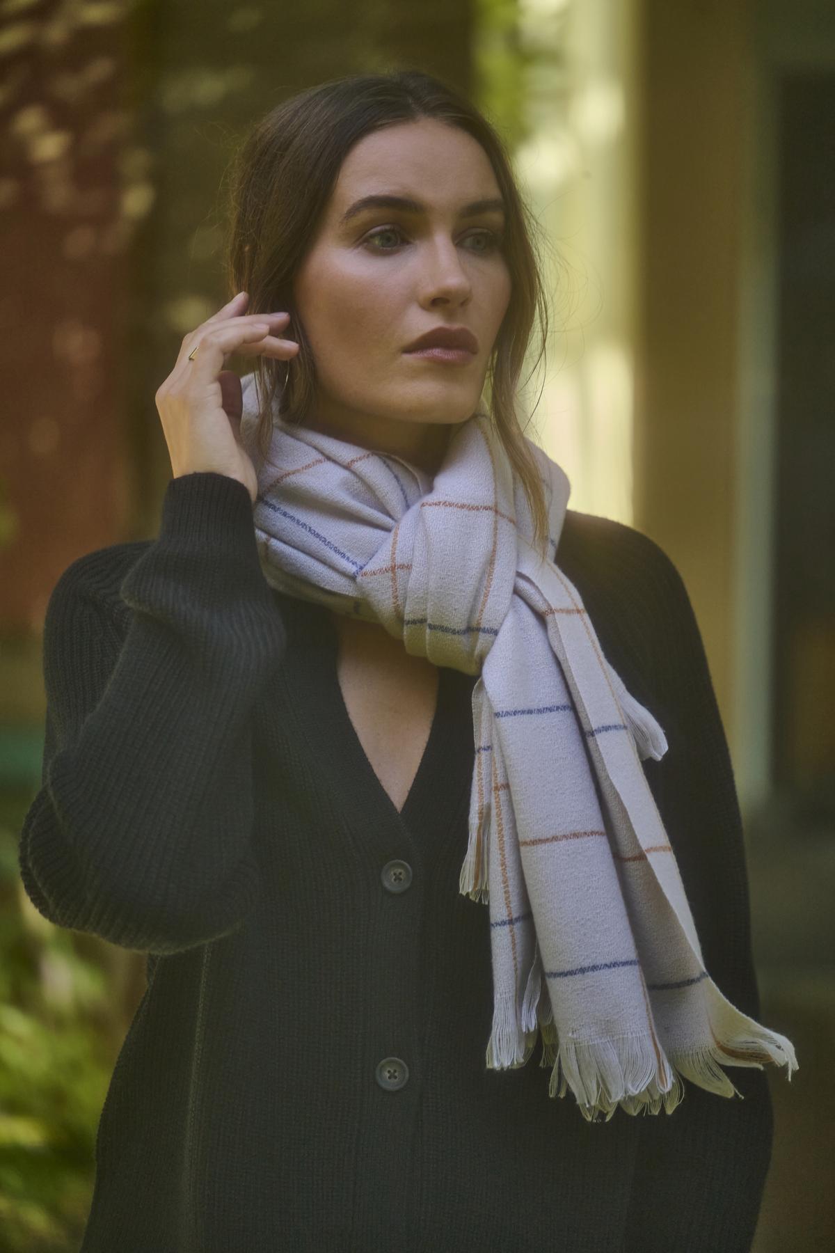   A person with long dark hair stands outdoors, wearing a black sweater and the HUDSON WINDOWPANE SCARF by Velvet by Graham & Spencer. They appear to be looking off camera. 