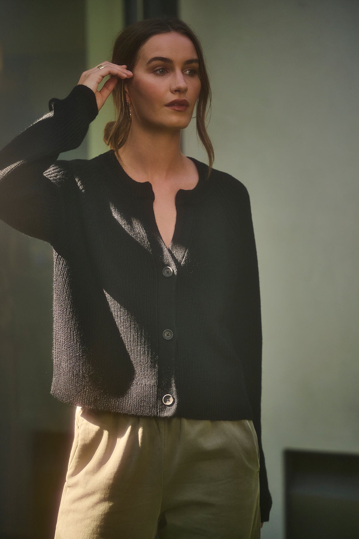 A person stands in a shadowed area, wearing the SHAYLA CARDIGAN by Velvet by Graham & Spencer—crafted from a cozy wool blend with raglan sleeves—and khaki pants, with one hand adjusting their hair.-38139286159553