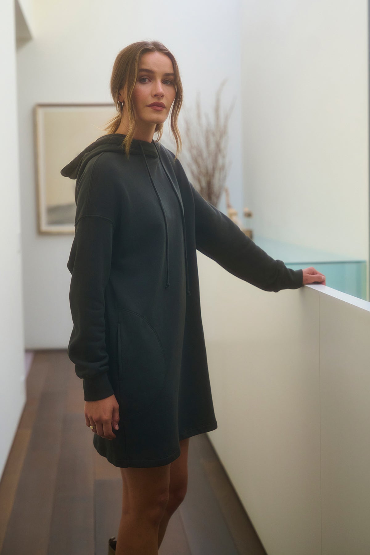 A person stands in a hallway wearing the ETTA HOODIE DRESS from Velvet by Graham & Spencer, casually leaning on a white railing. A framed picture and a vase of branches decorate the background.-38139294908609