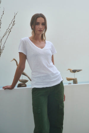 A woman wearing the Velvet by Graham & Spencer LILITH Tee and green cargo pants leans against a white surface, with decorative bird sculptures in the background.