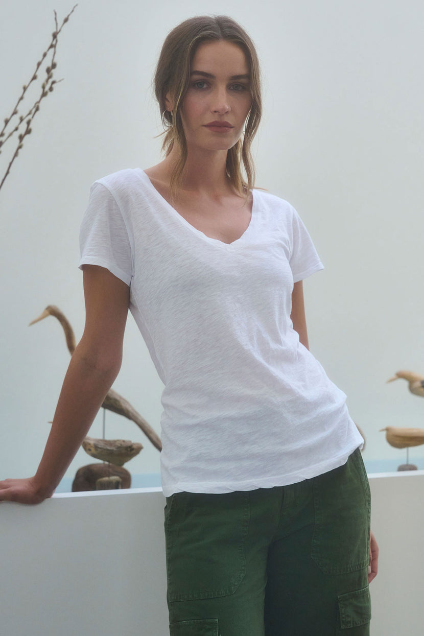 A woman wears the LILITH TEE by Velvet by Graham & Spencer—a white V-neck with a flattering fit—paired with green cargo pants, standing indoors near a white ledge featuring wooden bird decorations.