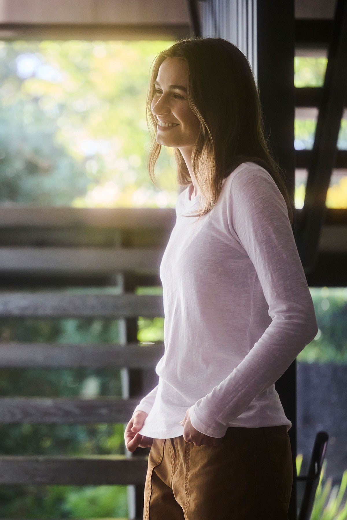   A woman in a LIZZIE TEE, a white crew-neck cotton slub shirt by Velvet by Graham & Spencer, paired with brown pants is smiling outdoors with her hands in her pockets. 