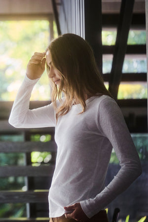 A woman stands indoors, wearing the LIZZIE TEE by Velvet by Graham & Spencer — a white cotton slub shirt with long sleeves and a crew neck — gently touching her hair as sunlight streams through the window.