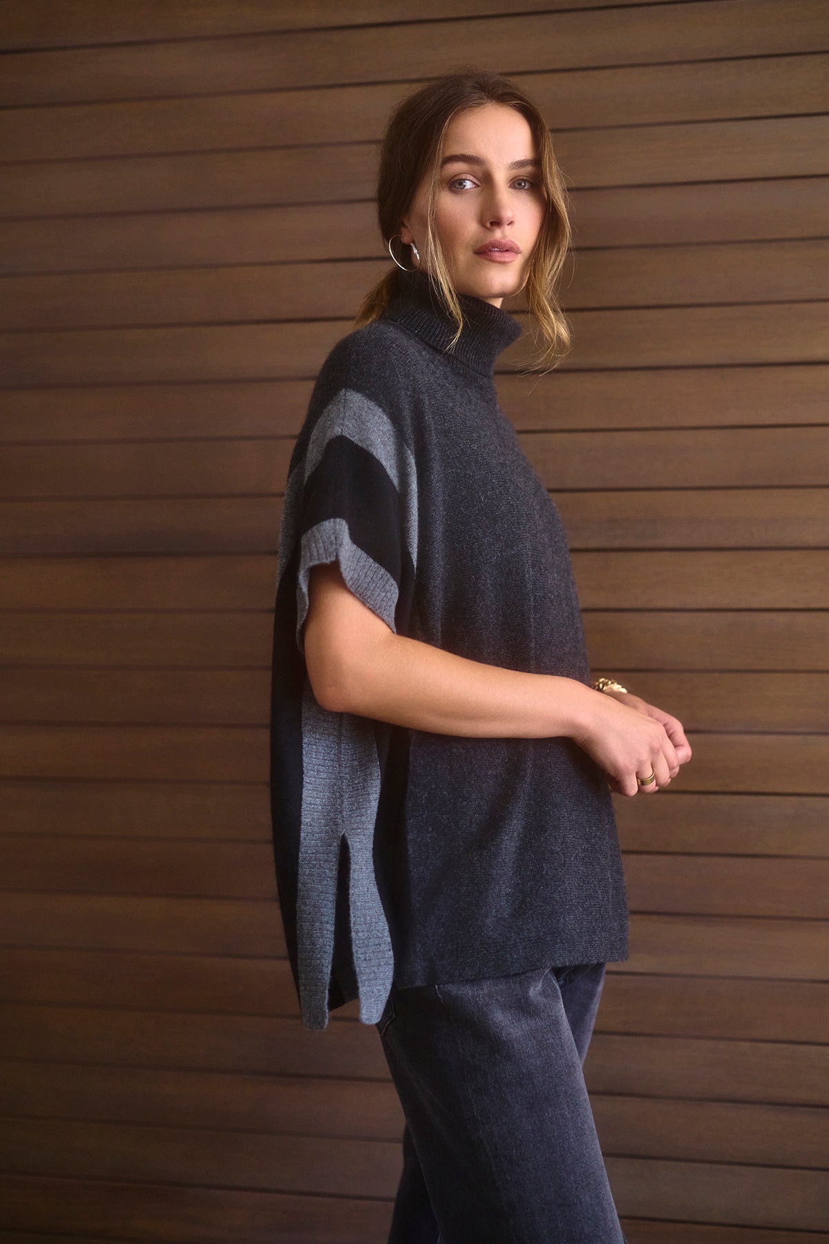   A person stands against a wooden wall, wearing the Velvet by Graham & Spencer REESE Cashmere Turtleneck Poncho in dark gray paired with gray pants, looking towards the camera. 