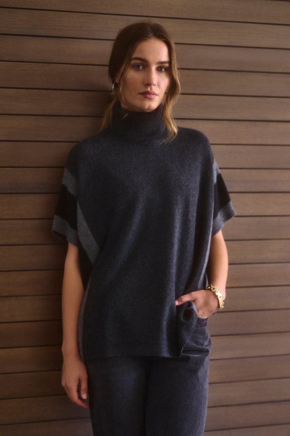   A woman in a Velvet by Graham & Spencer REESE CASHMERE TURTLENECK PONCHO and matching pants stands against a wooden wall, with one hand in her pocket, looking towards the camera. 