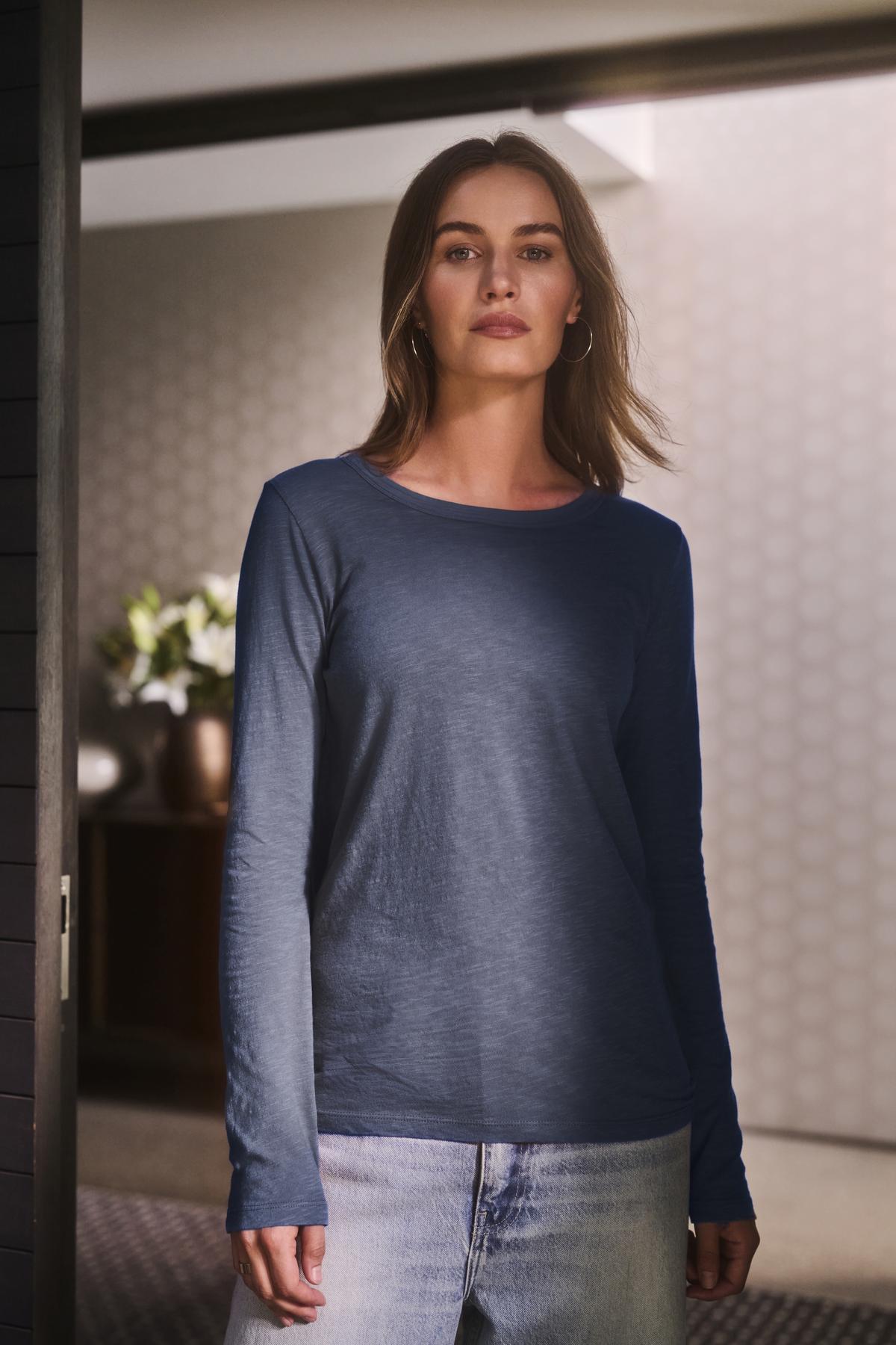   A person with shoulder-length hair wears the LIZZIE TEE by Velvet by Graham & Spencer, styled with light jeans while standing in a modern interior space. 