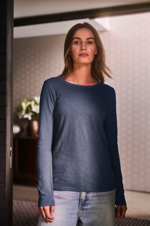 A person with shoulder-length hair wears the LIZZIE TEE by Velvet by Graham & Spencer, styled with light jeans while standing in a modern interior space.