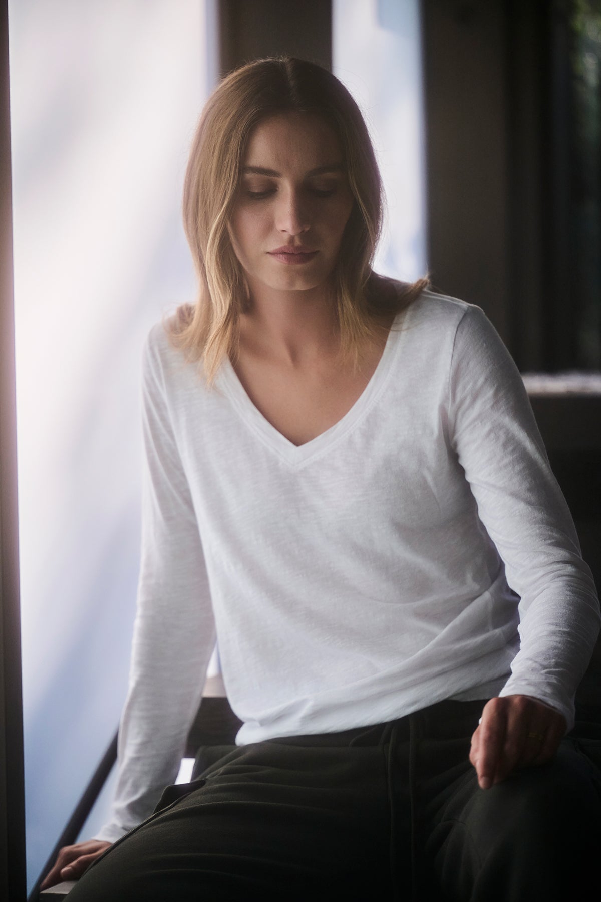   A person with shoulder-length hair, wearing the BLAIRE TEE from Velvet by Graham & Spencer, which features a white textured cotton slub and a v-neckline, is seated indoors and looking down. 