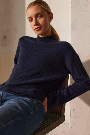 A person in a SIMONE CASHMERE MOCK NECK SWEATER by Velvet by Graham & Spencer and jeans sits on a chair, leaning slightly against a concrete wall.