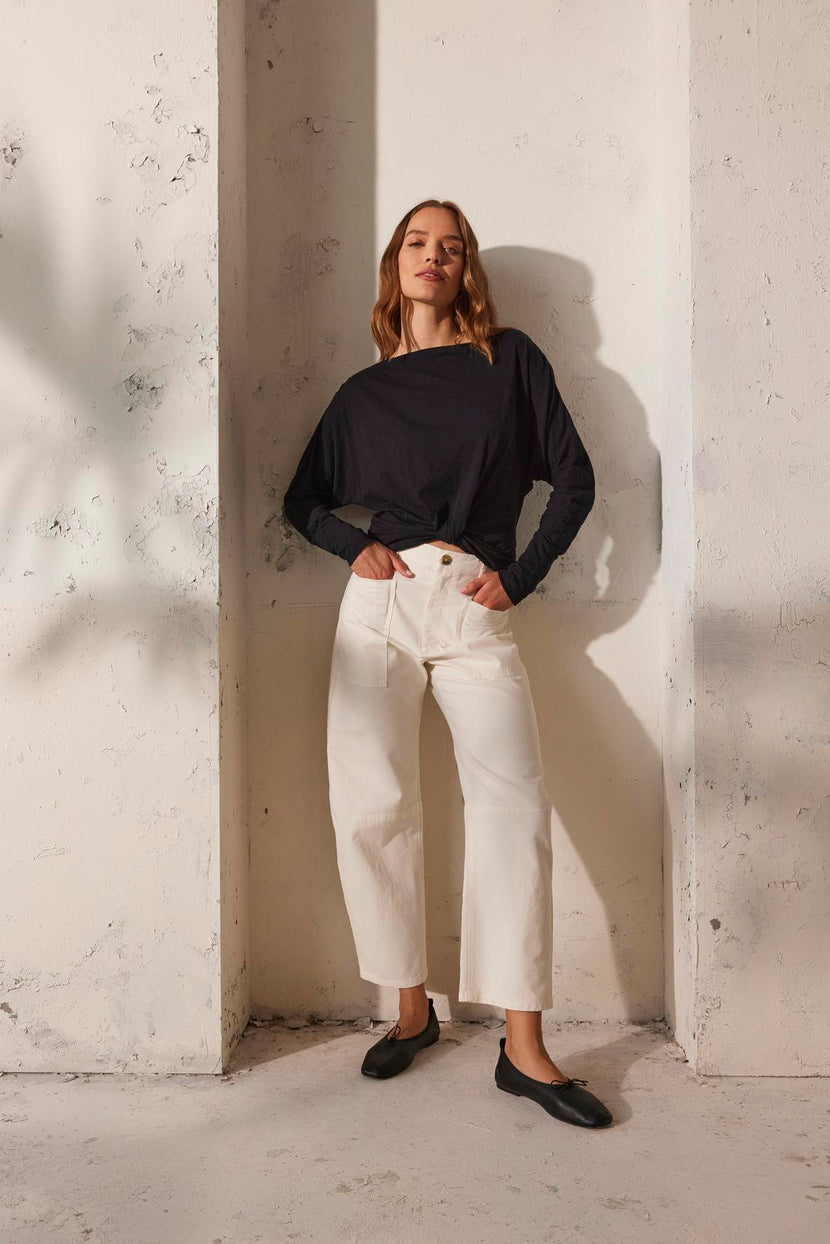 Against a textured white wall, a person wears a black long-sleeve shirt, the BRYLIE SANDED TWILL UTILITY PANT by Velvet by Graham & Spencer, and black shoes.