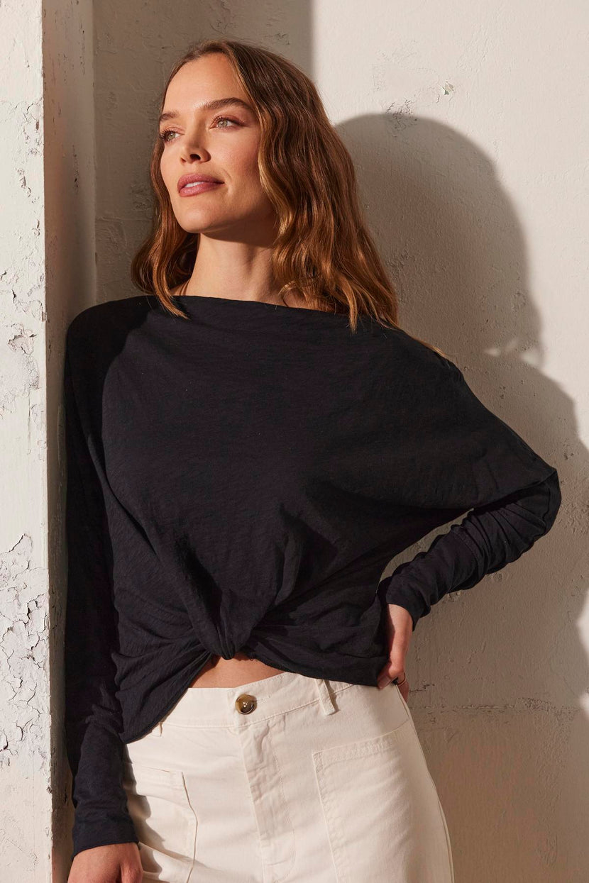 A woman with long hair leans against a textured white wall, wearing the HALLIE TEE by Velvet by Graham & Spencer, which has a black boat neckline and long sleeves, paired with white pants.