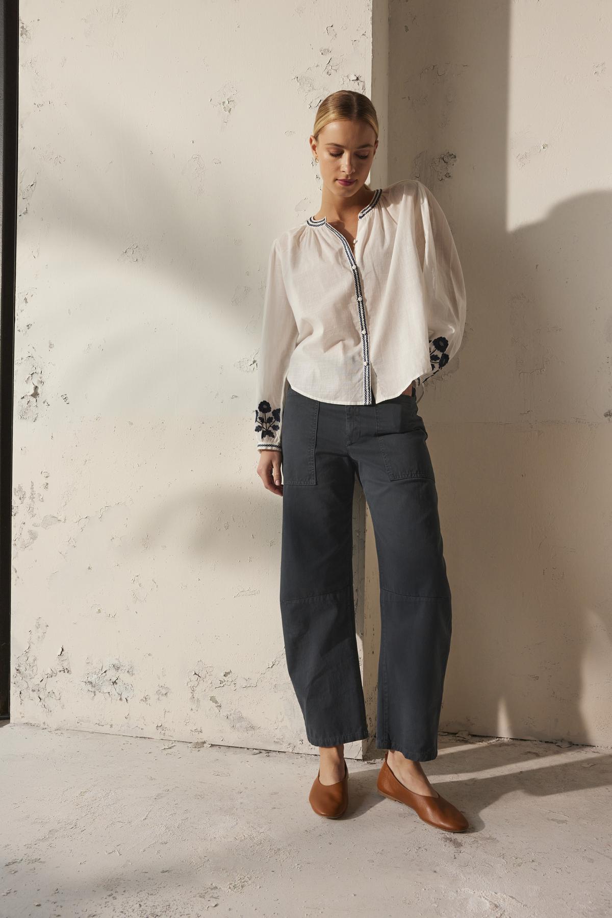 A person wearing the Velvet by Graham & Spencer BRYLIE SANDED TWILL UTILITY PANT and a white blouse stands against a textured wall, hands in the patch pockets.-38512701407425