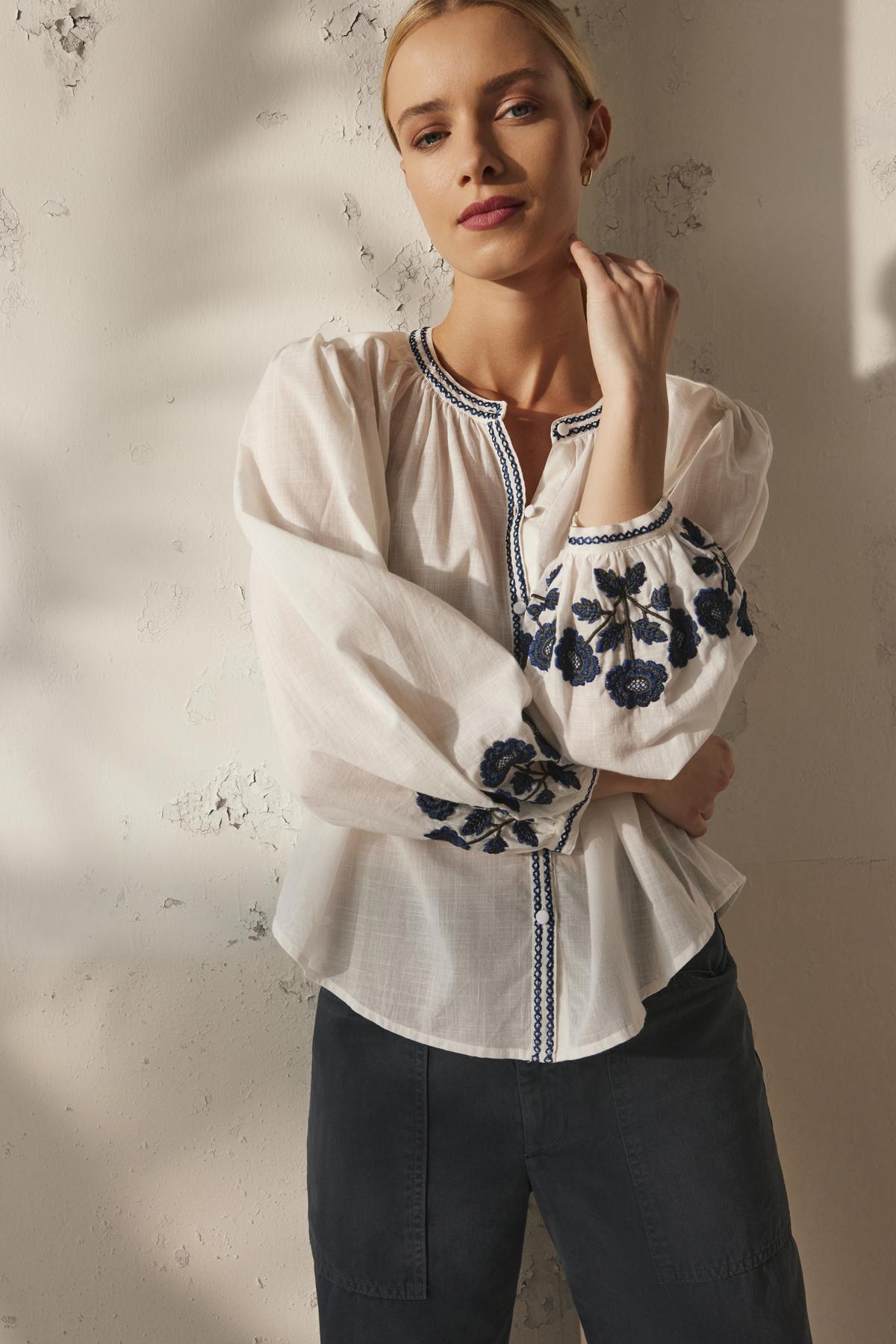   A person wearing the RAHWA TOP by Velvet by Graham & Spencer, a white cotton embroidered blouse with a button front, pairs it with dark pants and stands against a textured wall, looking at the camera. 