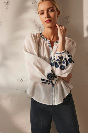 A person wearing the RAHWA TOP by Velvet by Graham & Spencer, a white cotton embroidered blouse with a button front, pairs it with dark pants and stands against a textured wall, looking at the camera.