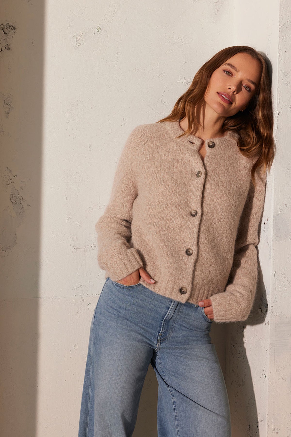   A person leans against a textured wall, wearing the CHANTAL ALPACA CARDIGAN from Velvet by Graham & Spencer paired with light blue jeans, with hands in pockets. 