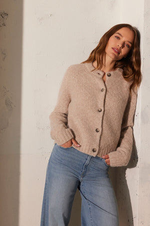 A person leans against a textured wall, wearing the CHANTAL ALPACA CARDIGAN from Velvet by Graham & Spencer paired with light blue jeans, with hands in pockets.