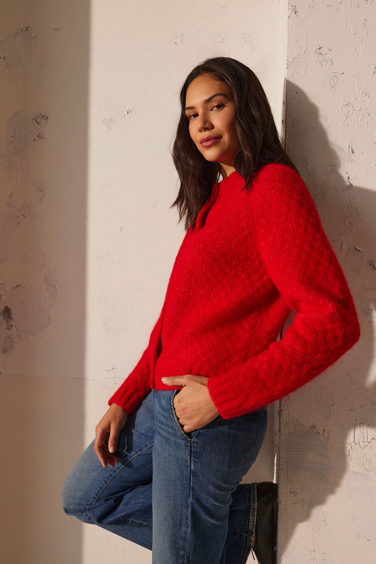   A person wearing the CHANTAL ALPACA CARDIGAN by Velvet by Graham & Spencer, paired with blue jeans, leans against a textured wall with one hand in their pocket. 