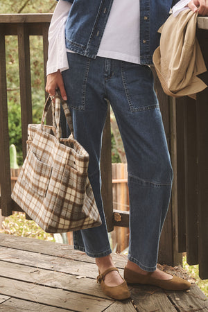 A person stands on a wooden deck wearing denim pants and vest, a white shirt, brown flats, holding the Velvet by Graham & Spencer PLAID TOTE crafted from a polyester wool blend, and carrying a beige jacket.