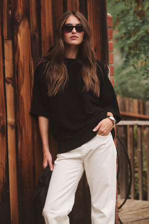 A woman wearing sunglasses and the COCO SWEATER by Velvet by Graham & Spencer over a black top with white pants leans against a wooden wall outdoors, holding a dark item in one hand.
