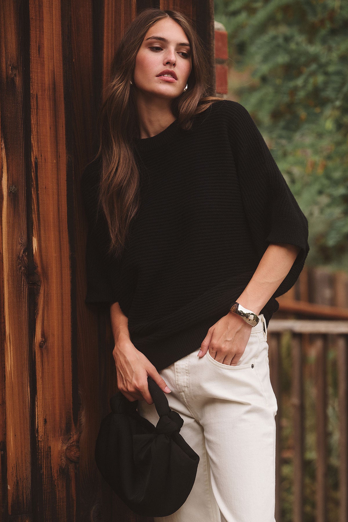 A woman stands against a wooden wall, wearing the COCO SWEATER by Velvet by Graham & Spencer in black with white pants, holding a black handbag; greenery and a fence are visible in the background.-39839138775233