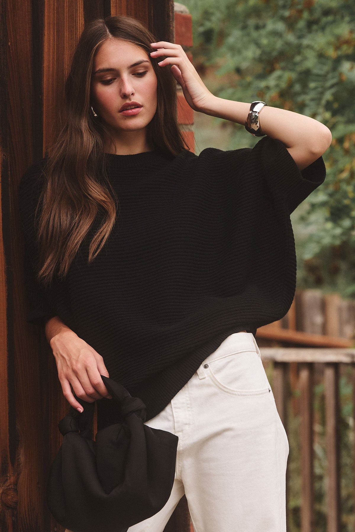 A woman stands outdoors against a wooden wall, wearing a loose black top and white pants, holding the ROBYN SATIN BAG by Velvet by Jenny Graham, with greenery in the background.-39839125143745