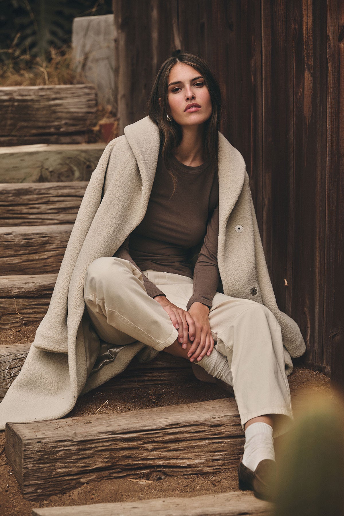   A woman sits on wooden steps outdoors, wearing the ELEANOR SHERPA REVERSIBLE COAT by Velvet by Graham & Spencer draped over her shoulders, paired with a brown top, light pants, white socks, and dark shoes. 