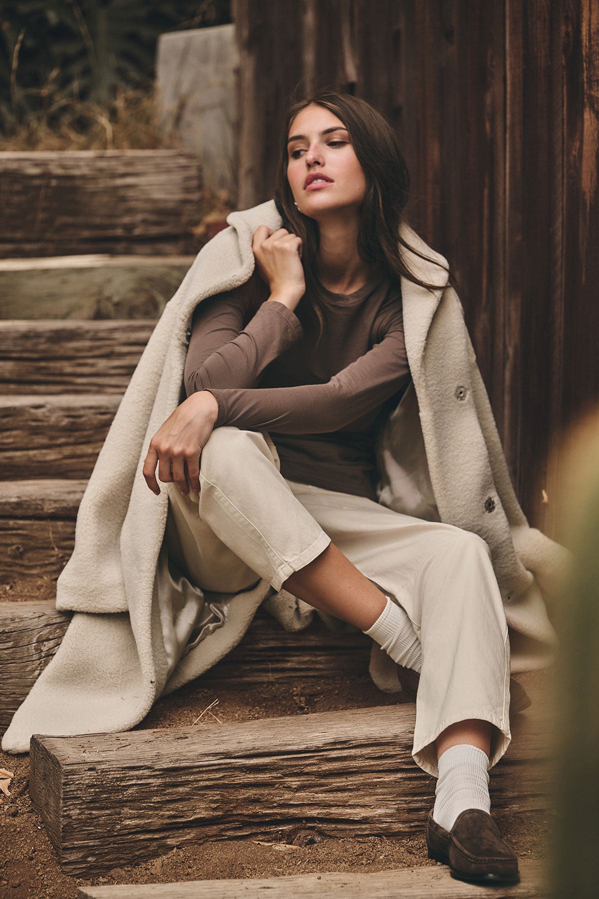 A woman sits on outdoor wooden steps wearing a long cream coat, a brown long-sleeve top, white socks, dark shoes, and the BRYLIE SANDED TWILL UTILITY PANT by Velvet by Graham & Spencer in cream.-39839382995137