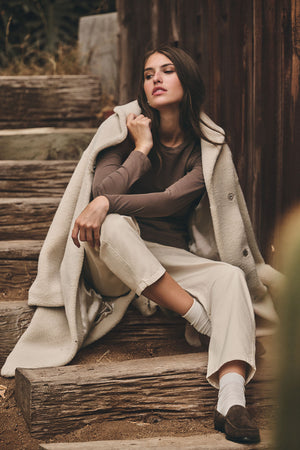 A woman sits on outdoor wooden steps wearing a long cream coat, a brown long-sleeve top, white socks, dark shoes, and the BRYLIE SANDED TWILL UTILITY PANT by Velvet by Graham & Spencer in cream.
