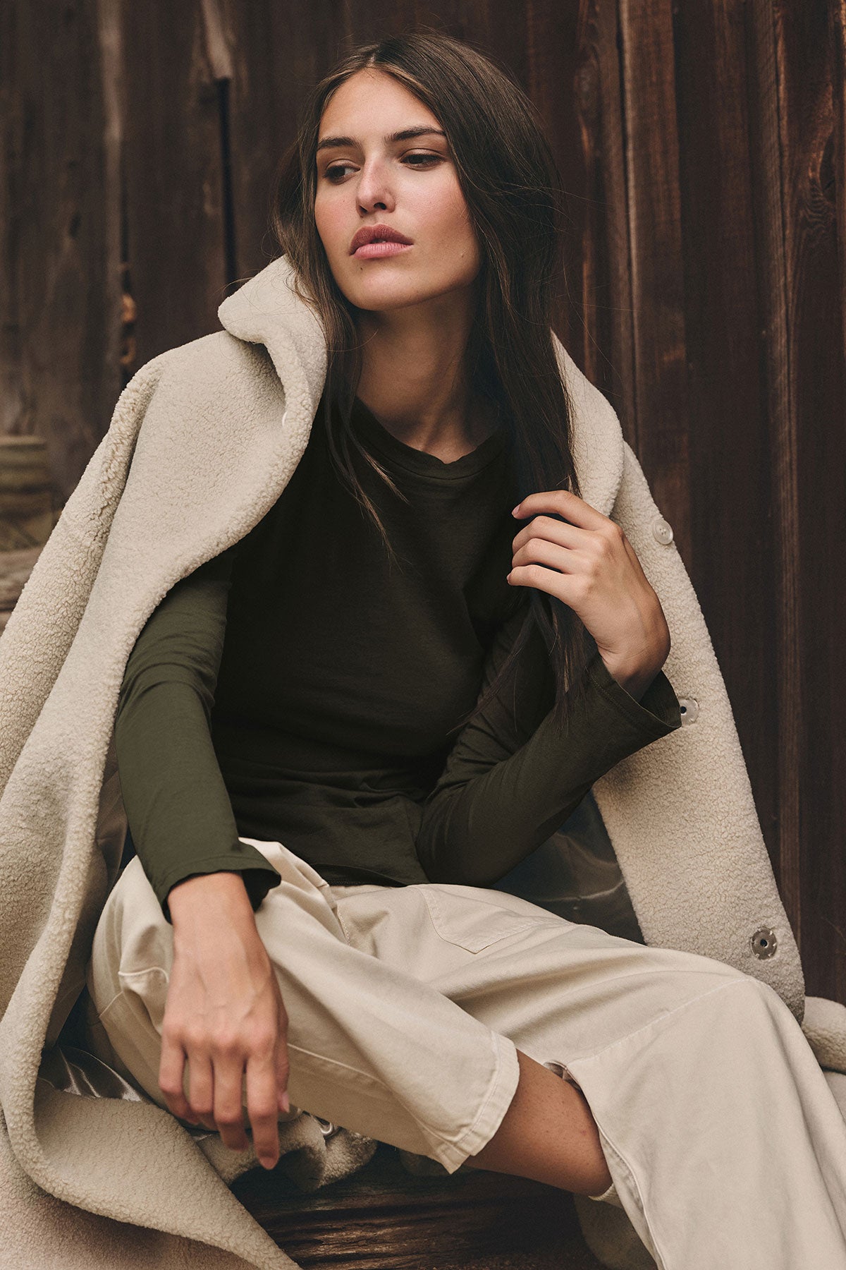   A woman sits outdoors wearing a light beige coat, beige pants, and the ZOFINA TEE by Velvet by Graham & Spencer—an ultra-soft, olive green fitted long sleeve crewneck—looking to the side with a neutral expression. 
