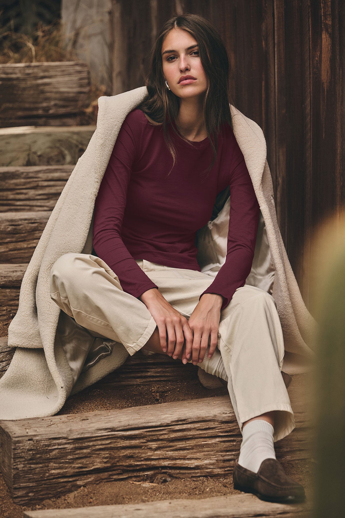 A woman sits on wooden steps wearing the ZOFINA TEE by Velvet by Graham & Spencer, cream pants, a light draped coat, white socks, and brown shoes, looking at the camera with a neutral expression.-40131025109185