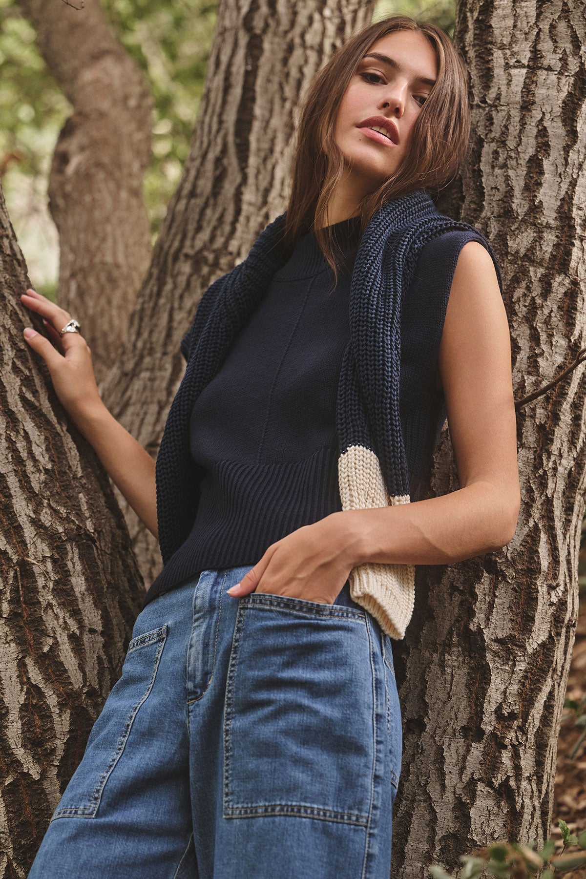   A woman stands outdoors by a large tree, wearing Velvet by Graham & Spencer's ELECTRA COTTON CASHMERE SWEATER VEST with ribbed hems, blue jeans, and a navy and cream knit scarf draped around her neck. 
