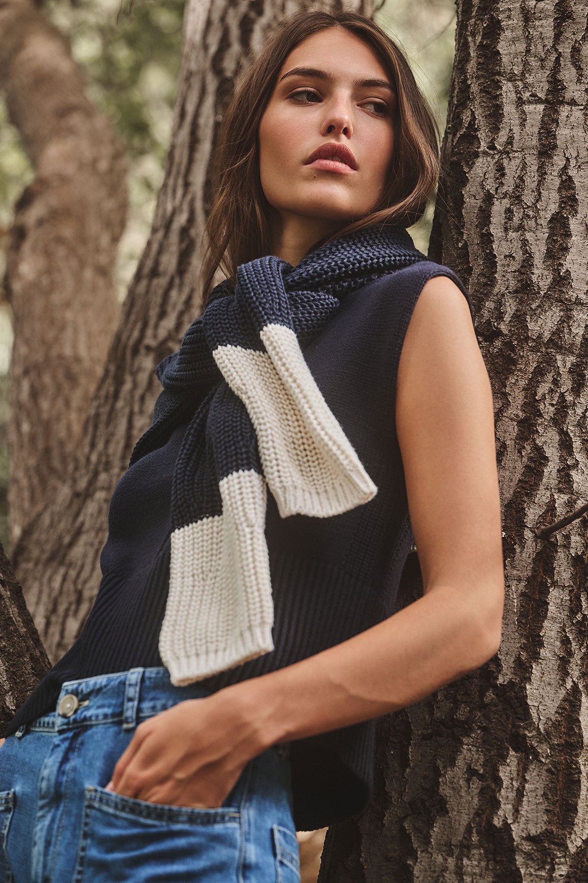   A woman in the SHAY SWEATER by Velvet by Graham & Spencer and blue jeans leans against a tree, adding a navy and white knit scarf for cozy layering. 