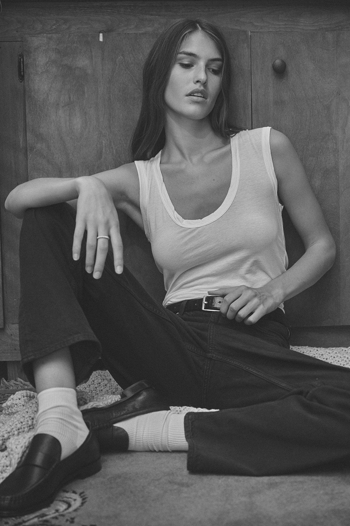 Woman sits on floor wearing Velvet by Graham & Spencer’s ESTINA TANK TOP with dark pants, socks, and loafers; her arm rests on her knee. Black and white photo shows a neutral expression.-39840680902849