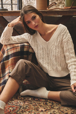 A woman wearing the JOYCE SWEATER by Velvet by Graham & Spencer and brown pants sits on a patterned rug, leaning against a plaid blanket draped over a chair.