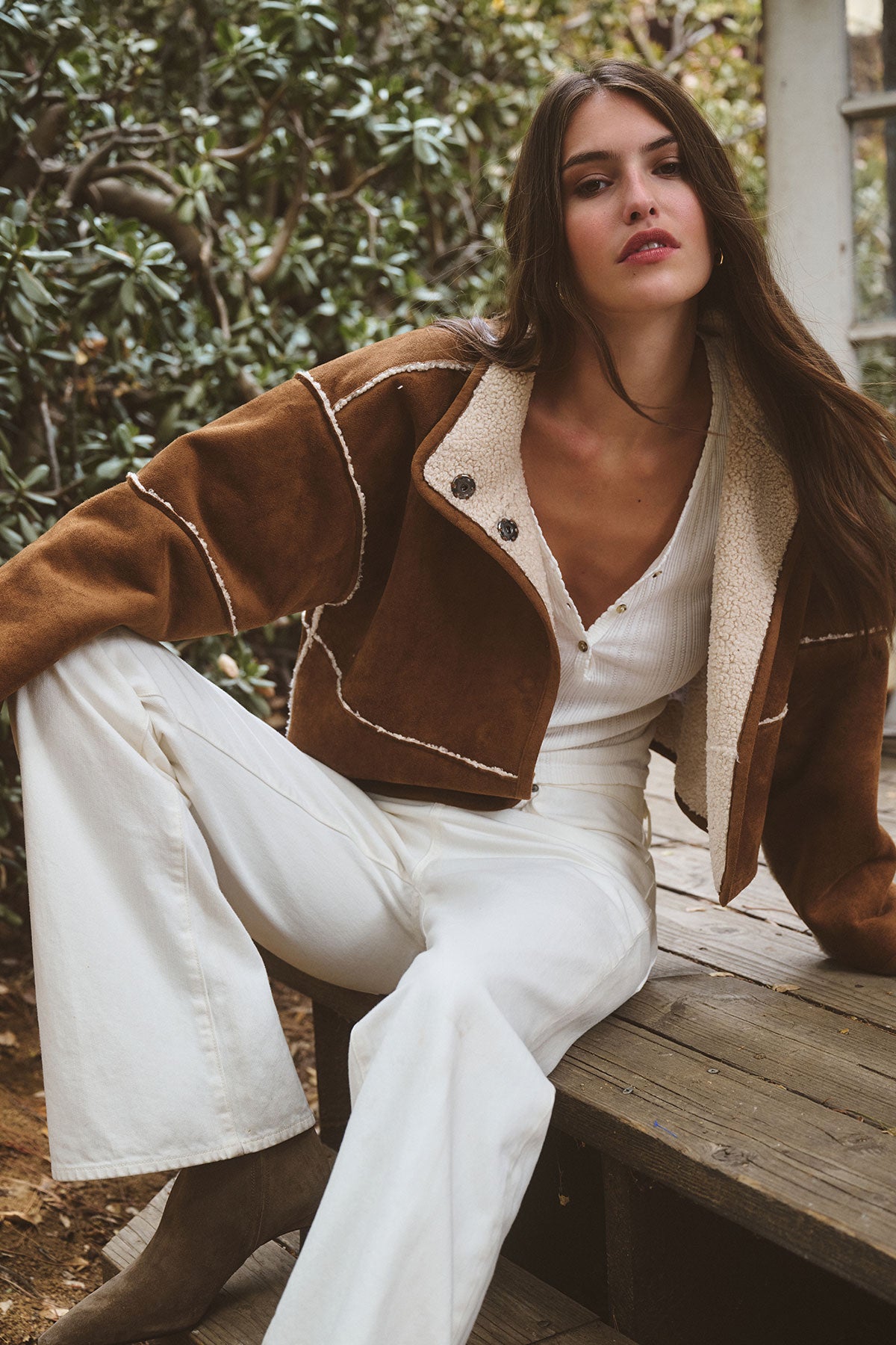 A woman sits on wooden steps outdoors, wearing the Velvet by Graham & Spencer KELLY LUXE SHERPA REVERSIBLE JACKET over a white buttoned jumpsuit, with greenery in the background.-39839522750657