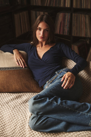A woman wearing the Velvet by Graham & Spencer RAK TEE and jeans reclines on a beige couch with a brown cushion, with a bookshelf of records in the background.