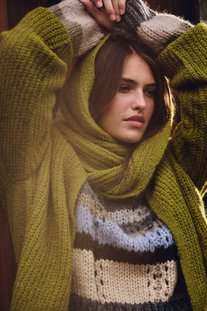 A woman in a green LUCA ALPACA SCARF by Velvet by Graham & Spencer and a cardigan over a multicolored striped sweater stands with her arms raised above her head.