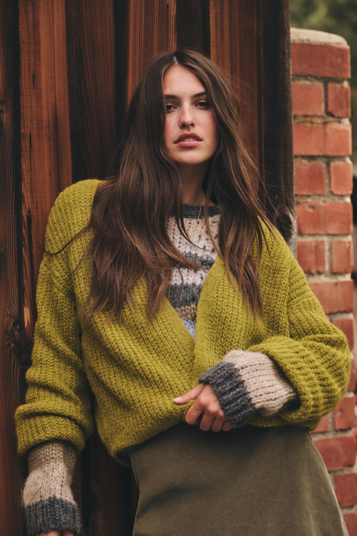 A woman with long brown hair wears the RANYA ALPACA CARDIGAN by Velvet by Graham & Spencer over a patterned sweater, standing against a wooden wall and brick column.-39850588045505