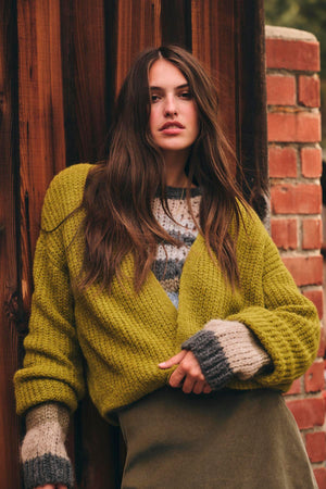 A woman with long brown hair wears the RANYA ALPACA CARDIGAN by Velvet by Graham & Spencer over a patterned sweater, standing against a wooden wall and brick column.